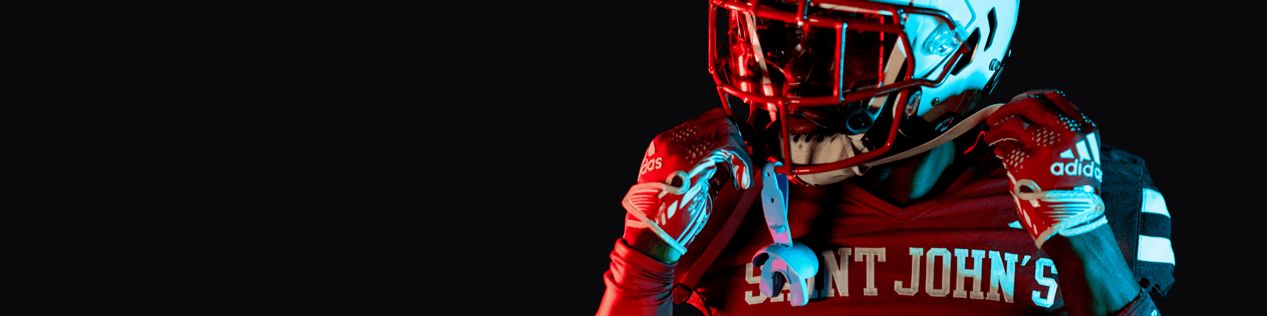 A football player in a red "Saint John's" jersey and helmet stands against a black background, illuminated by dramatic red and blue lighting, holding his helmet strap with both hands.
