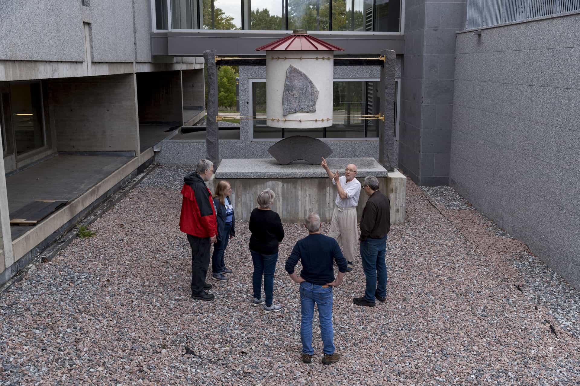 A group of six people stands on gravel, listening to a man in white who gestures toward a large stone slab displayed on a structure between modern concrete buildings.