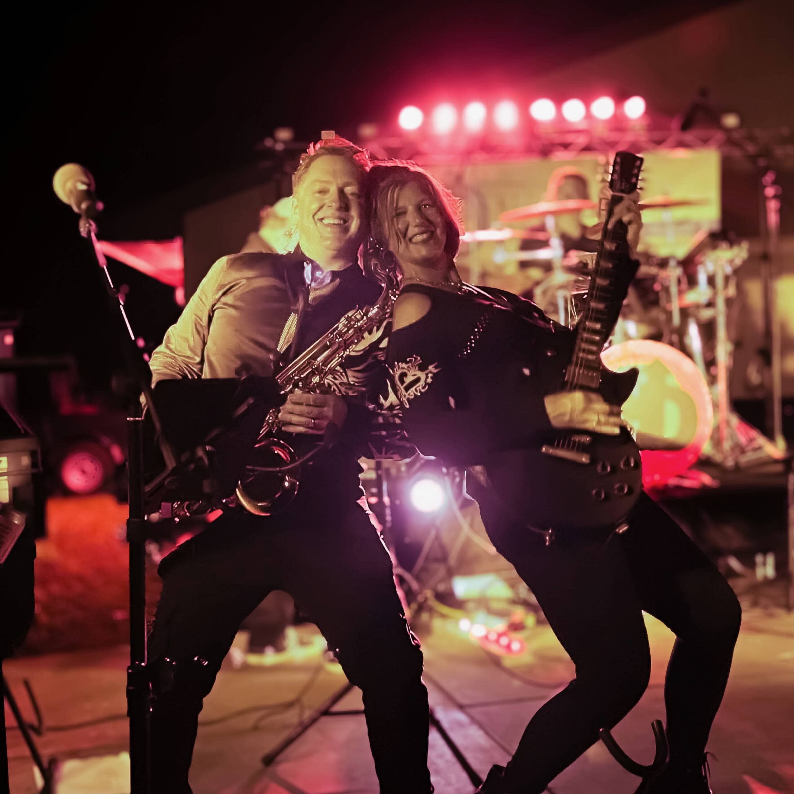 Two musicians, one playing saxophone and the other playing electric guitar, perform energetically onstage under bright, colorful lights, smiling and leaning close together. Drums and more lights are visible in the background.
