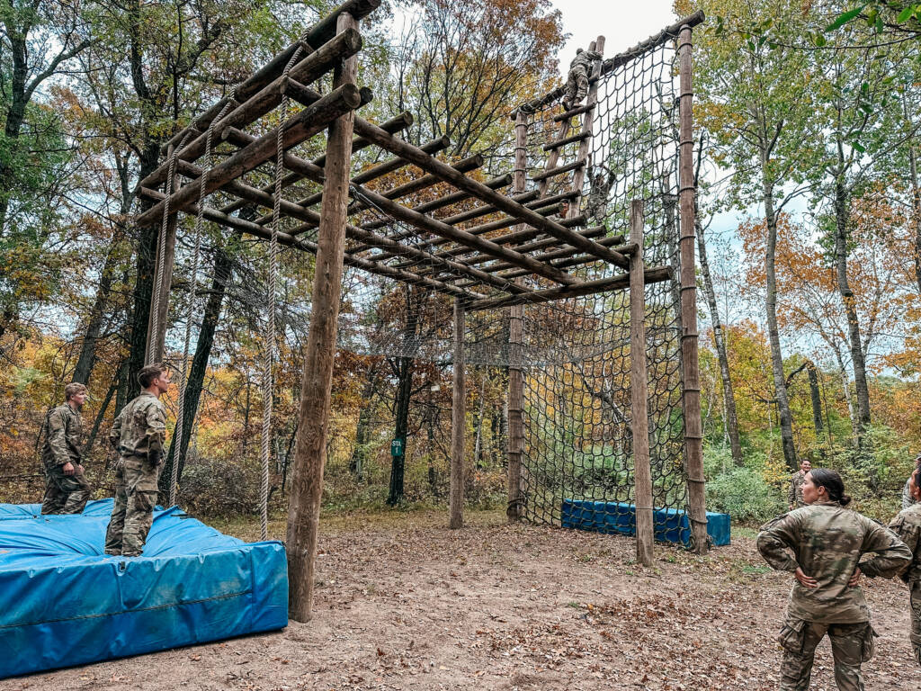 Several people in military uniforms observe a tall wooden obstacle course structure with rope nets and ladders, surrounded by trees and blue safety mats on the ground. The scene takes place outdoors in a wooded area.