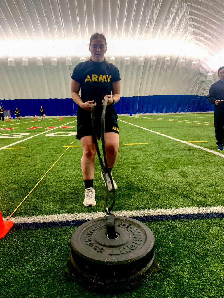 A person in an "ARMY" t-shirt and shorts pulls a weighted sled across an indoor turf field, participating in a fitness test. The background shows more people and orange cones under a large, domed ceiling.