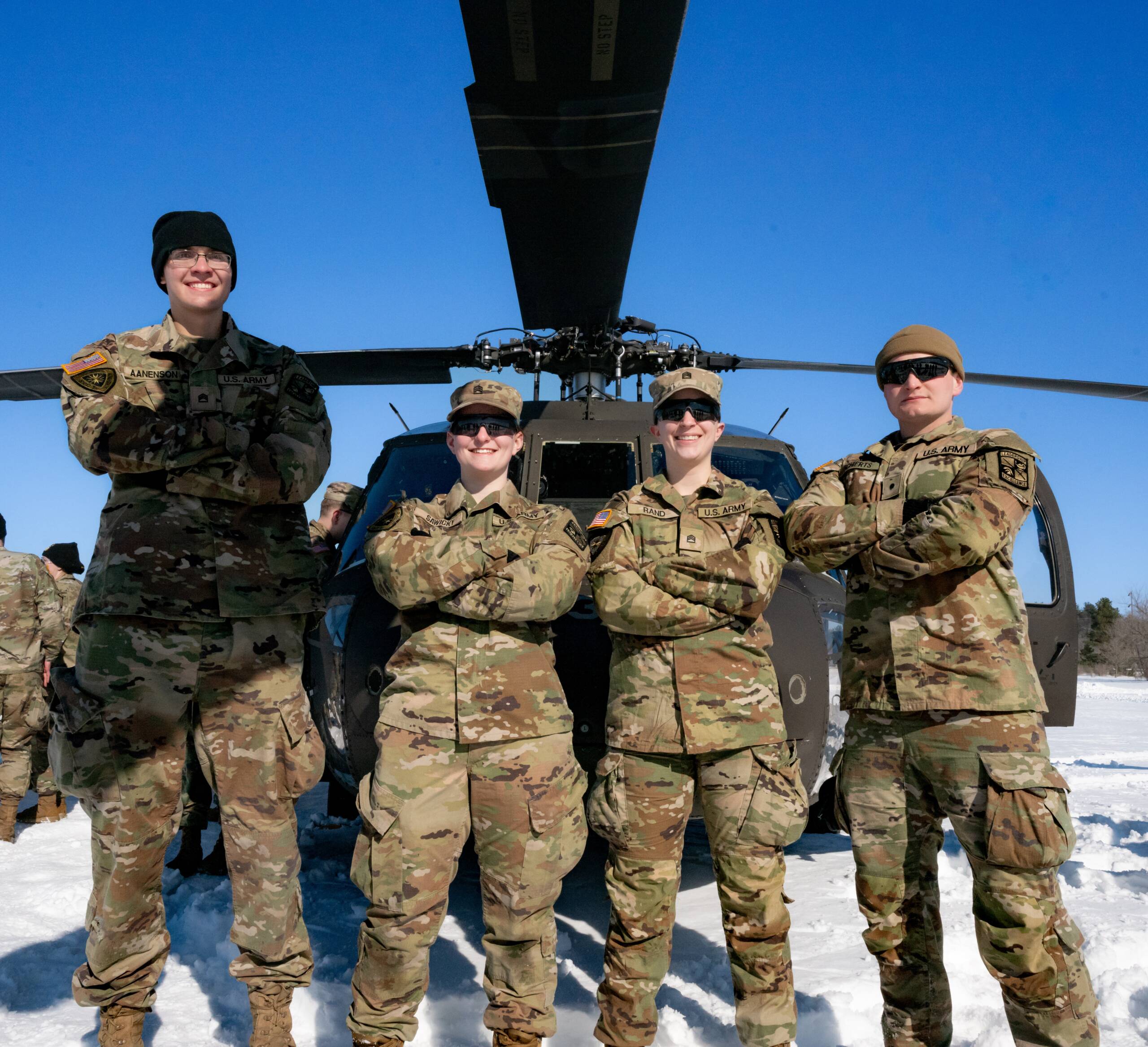Four soldiers in camouflage uniforms stand confidently with arms crossed in front of a military helicopter on snow-covered ground under a clear blue sky.