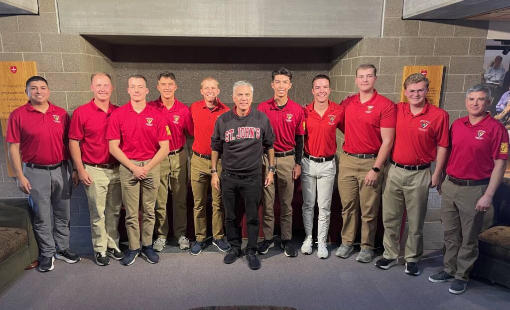 Ten men stand indoors, nine wearing matching red "St. John’s" polo shirts and khaki pants, and one in the center wearing a black "St. John’s" sweatshirt and black pants, smiling at the camera.