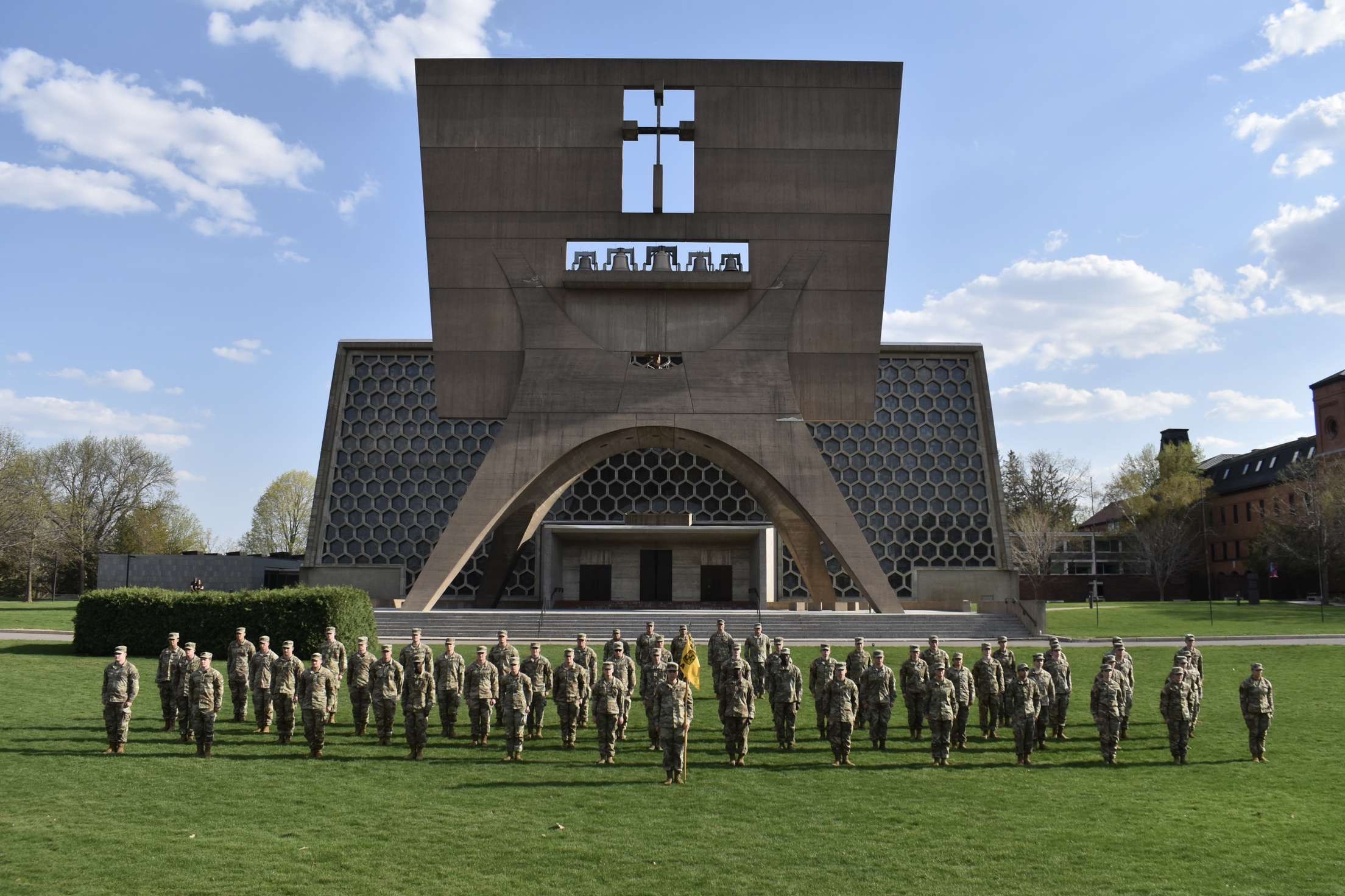 A group of uniformed soldiers stands in formation on a green lawn in front of a modern church building with a large cross and geometric patterns on its facade. The sky is partly cloudy.