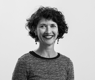 Black and white photo of a woman with short, curly hair, wearing a long-sleeve shirt and earrings. She is smiling and looking slightly to her left against a plain background.