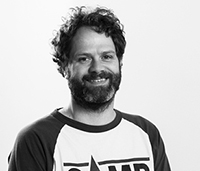 Black and white photo of a smiling person with curly hair and a beard, wearing a baseball-style shirt with contrasting sleeves and the word "CAMP" partially visible on the front. The background is a plain, light color.