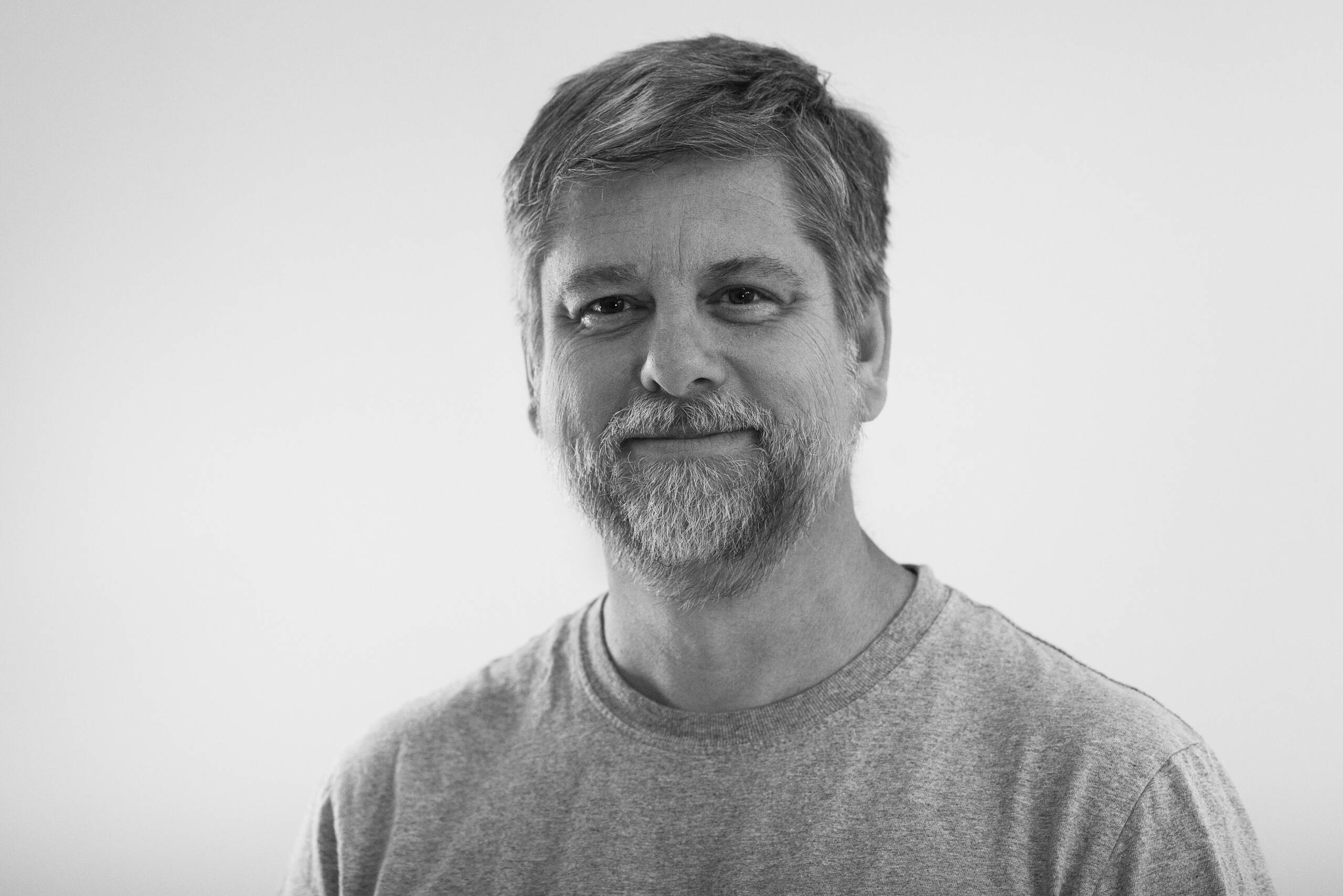 A middle-aged man with short hair and a beard, wearing a plain gray t-shirt, smiles slightly while looking at the camera. The background is plain and light-colored.