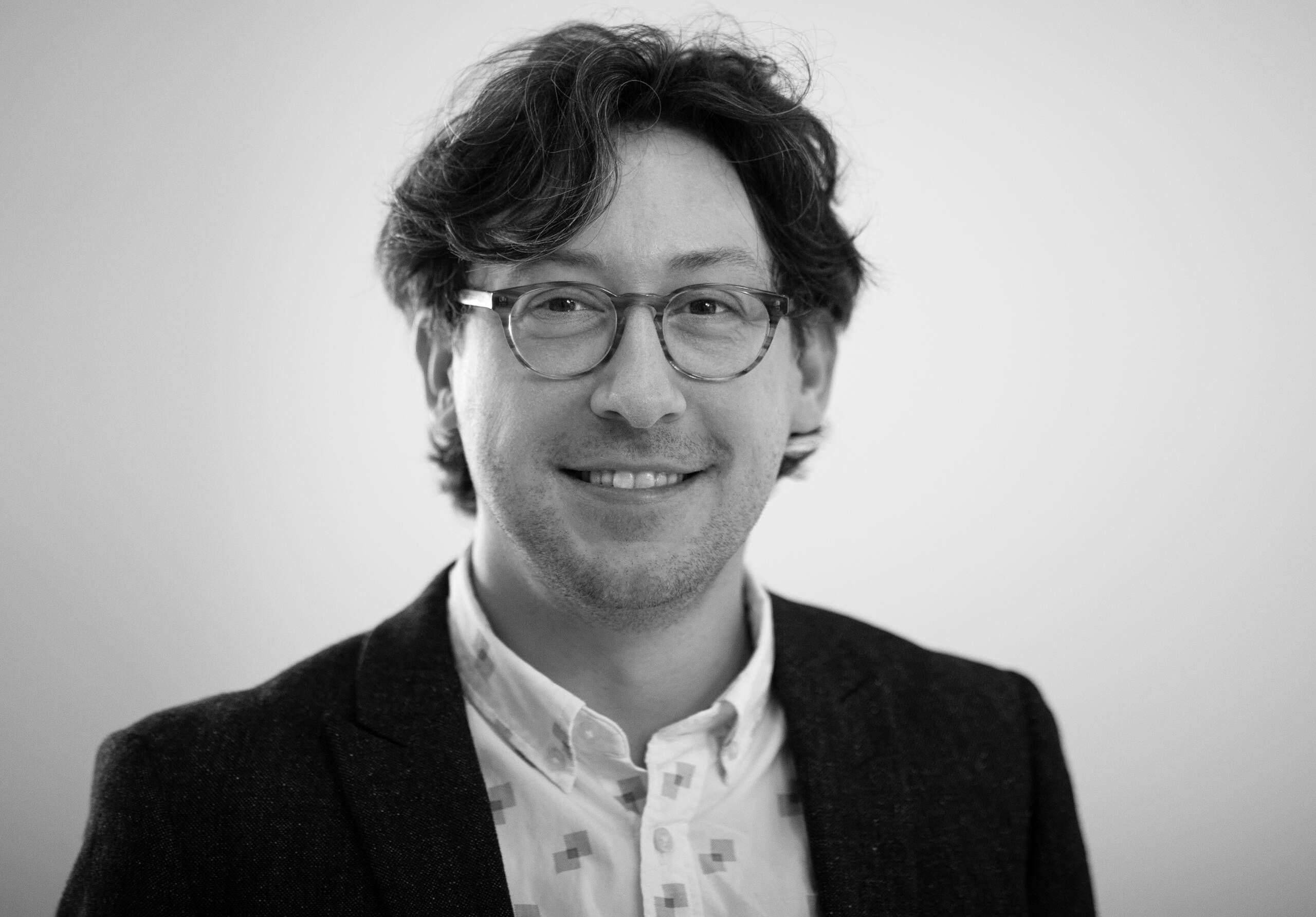A man with wavy dark hair, glasses, and a slight smile wears a patterned white shirt and a dark blazer. The background is plain and out of focus. The image is in black and white.