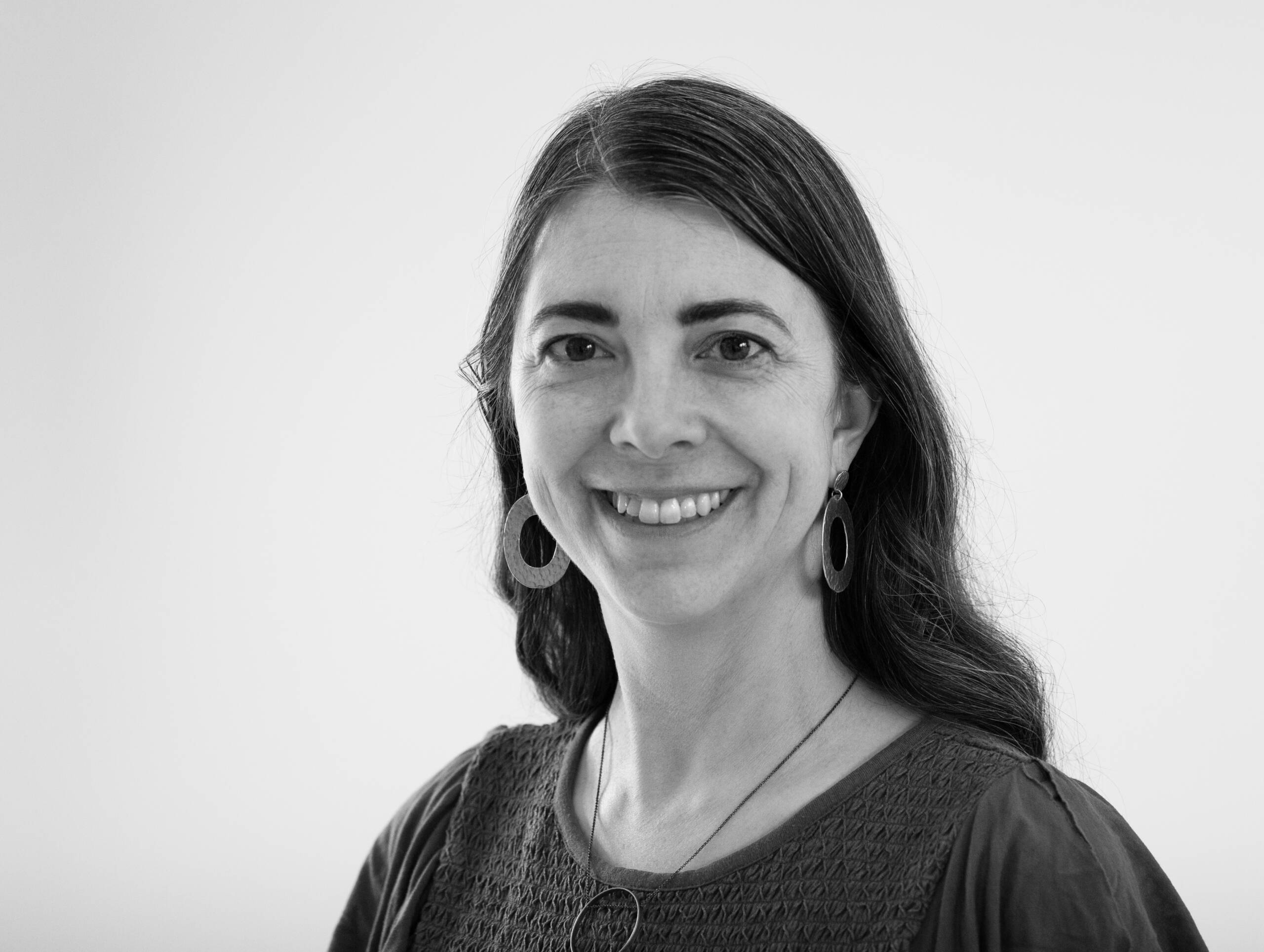 Black and white portrait of a woman with long dark hair, wearing circular earrings, a textured top, and a necklace, smiling and looking at the camera against a plain background.