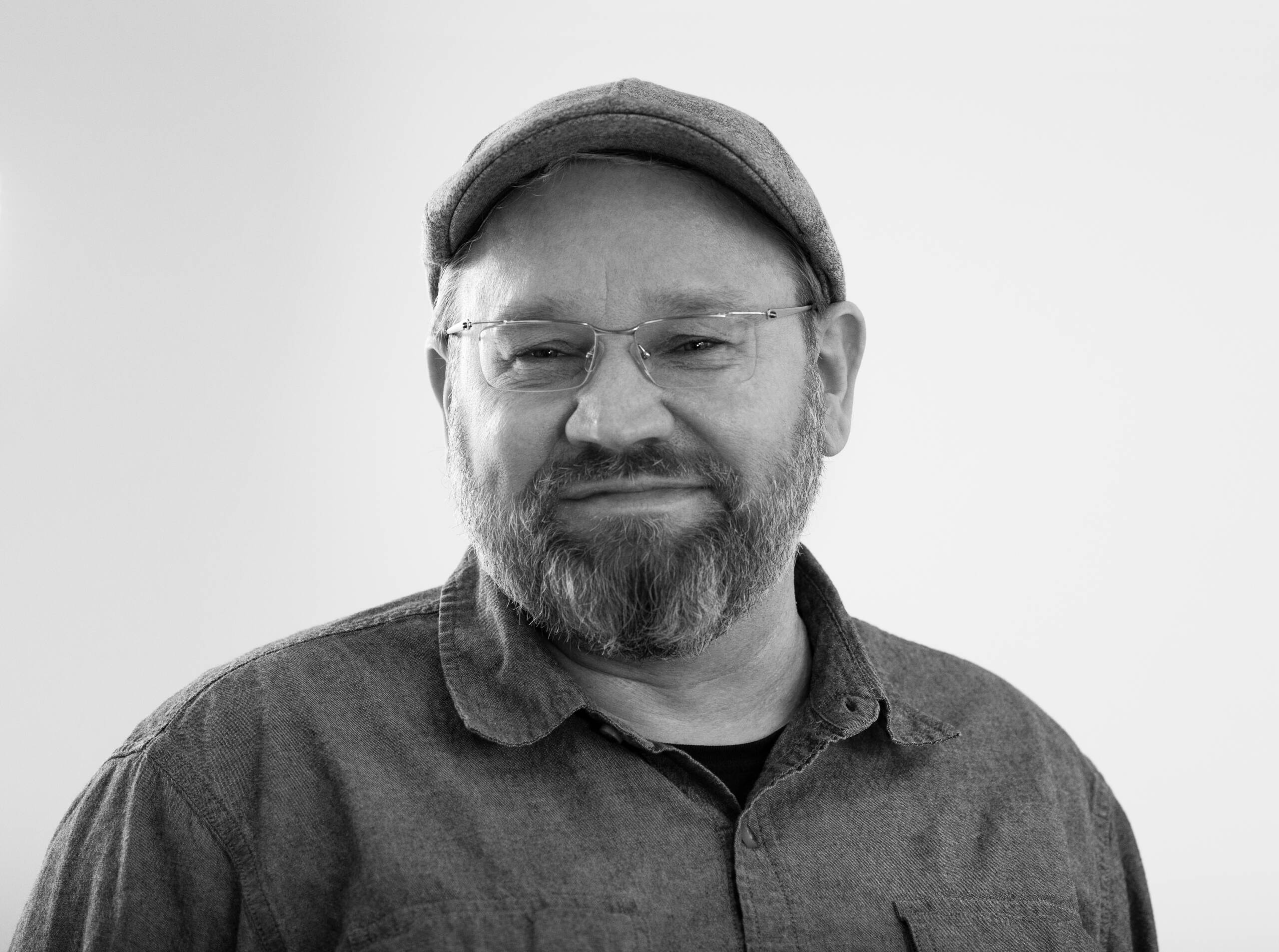 A man with a beard and glasses wearing a cap and a button-up shirt smiles slightly while facing the camera, set against a plain, light background.