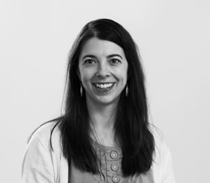 Black and white portrait of a woman with long, straight hair, wearing earrings, a button-up top, and a light-colored cardigan. She is smiling and looking directly at the camera with a plain background behind her.