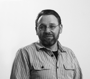 Black-and-white photo of a middle-aged man with short hair, wearing glasses and a striped button-up shirt with two breast pockets. He has a beard and is facing the camera with a slight smile, standing against a plain background.
