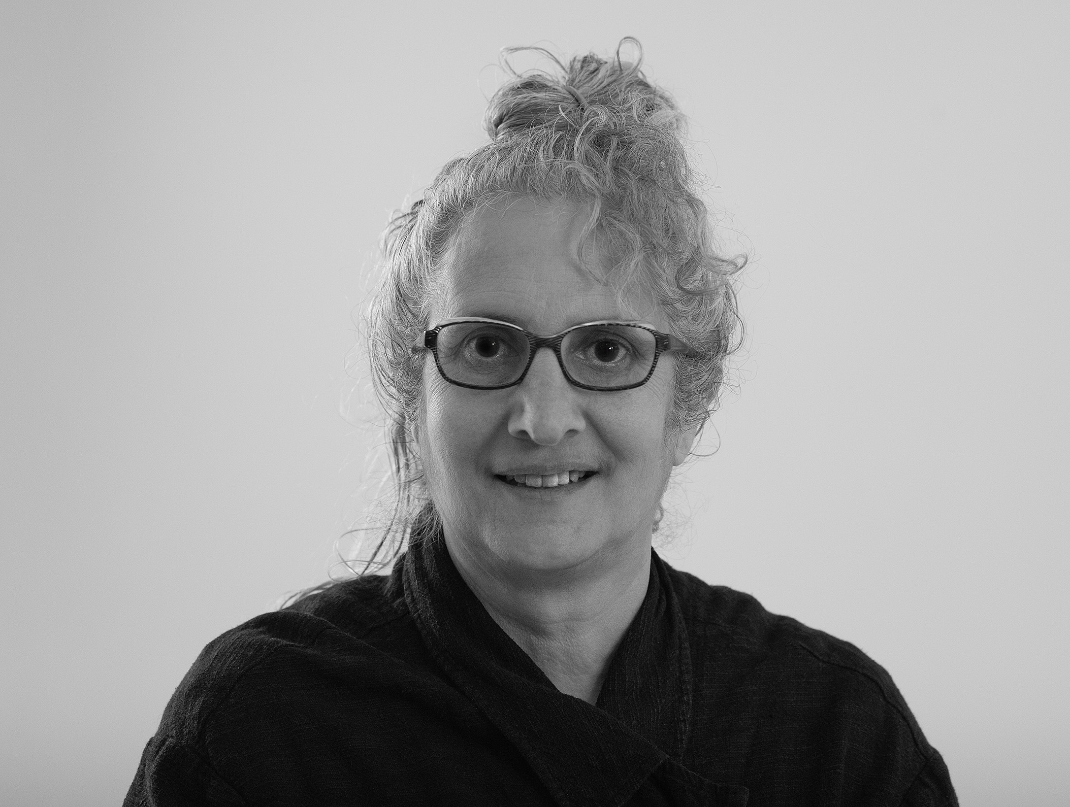 Black and white portrait of a person with curly hair tied up, wearing glasses and a dark textured top, smiling gently while looking at the camera against a plain background.