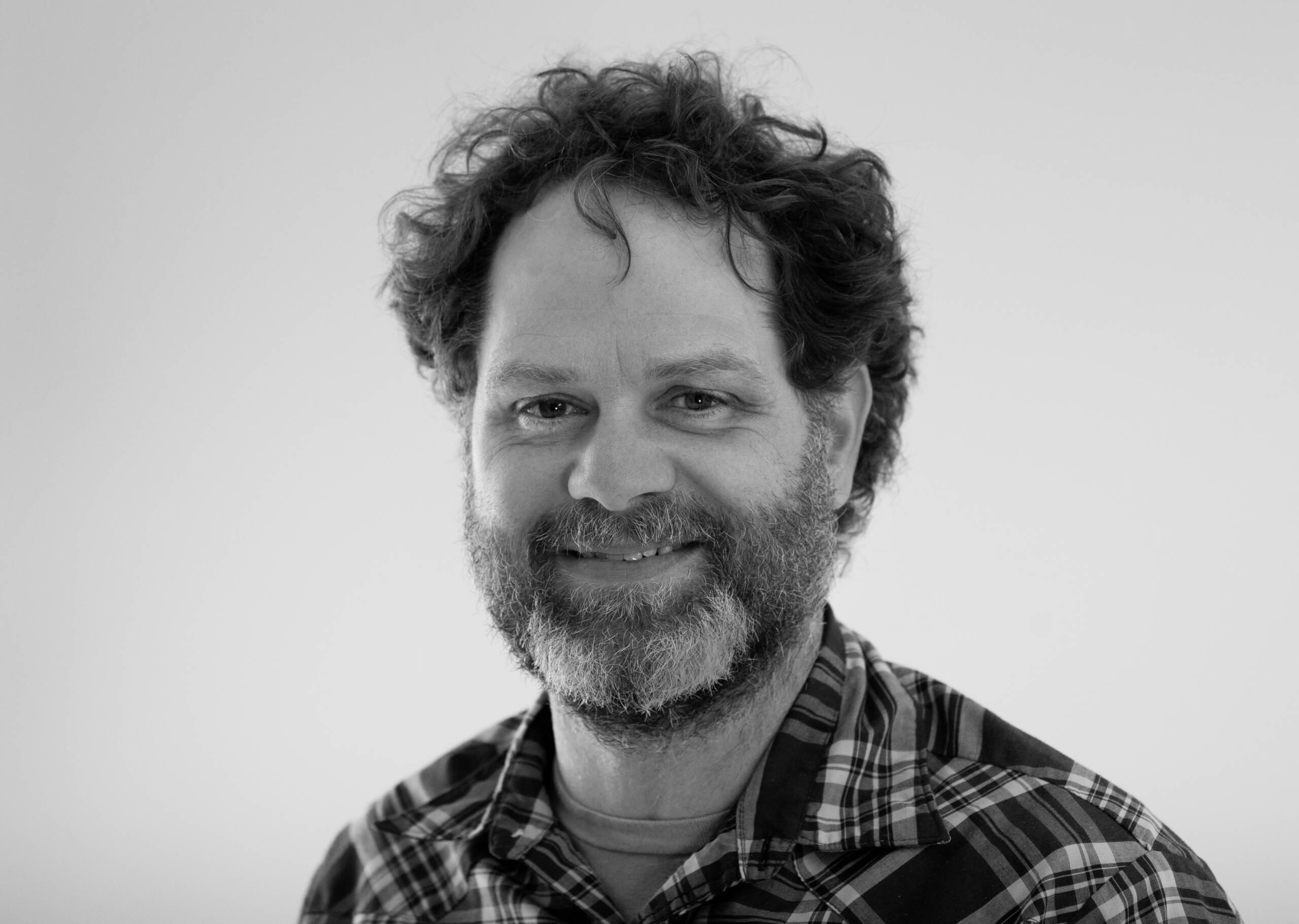 A man with curly hair and a beard smiles at the camera. He is wearing a plaid shirt and the background is plain and light-colored. The photo is black and white.