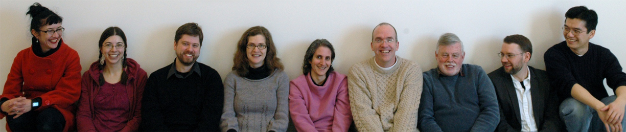 A group of eight people, six men and two women, sitting on a bench against a light-colored wall. They are all smiling and casually dressed in winter clothing, including sweaters and cardigans. They appear to be posing for a group photo.