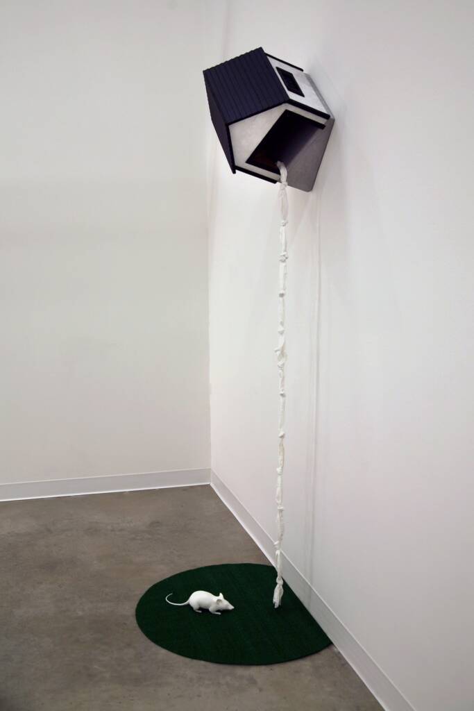 A small house sculpture is mounted high on a corner wall, with a rope made of tied fabric hanging down to a green mat below. A white mouse figurine sits on the mat, facing the rope.