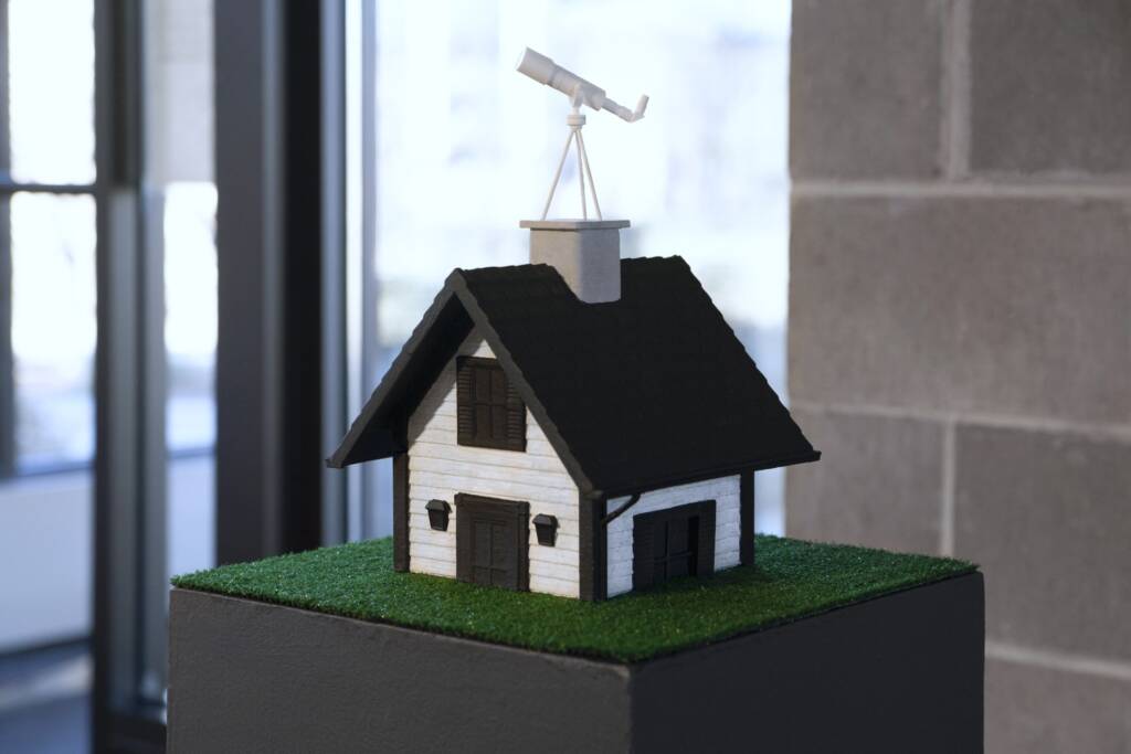 A small model of a white house with black shutters and roof sits on a patch of artificial grass, featuring a white telescope mounted on the chimney. The house is displayed indoors near a window and a brick wall.