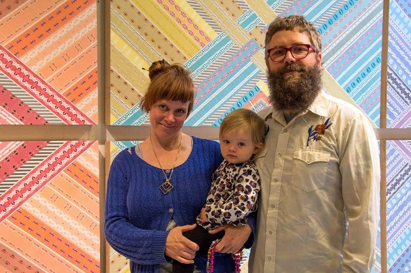 A woman, man, and child are standing together in front of a colorful, geometric-patterned wall. The woman holds the child, and the man stands beside them. All are looking at the camera.