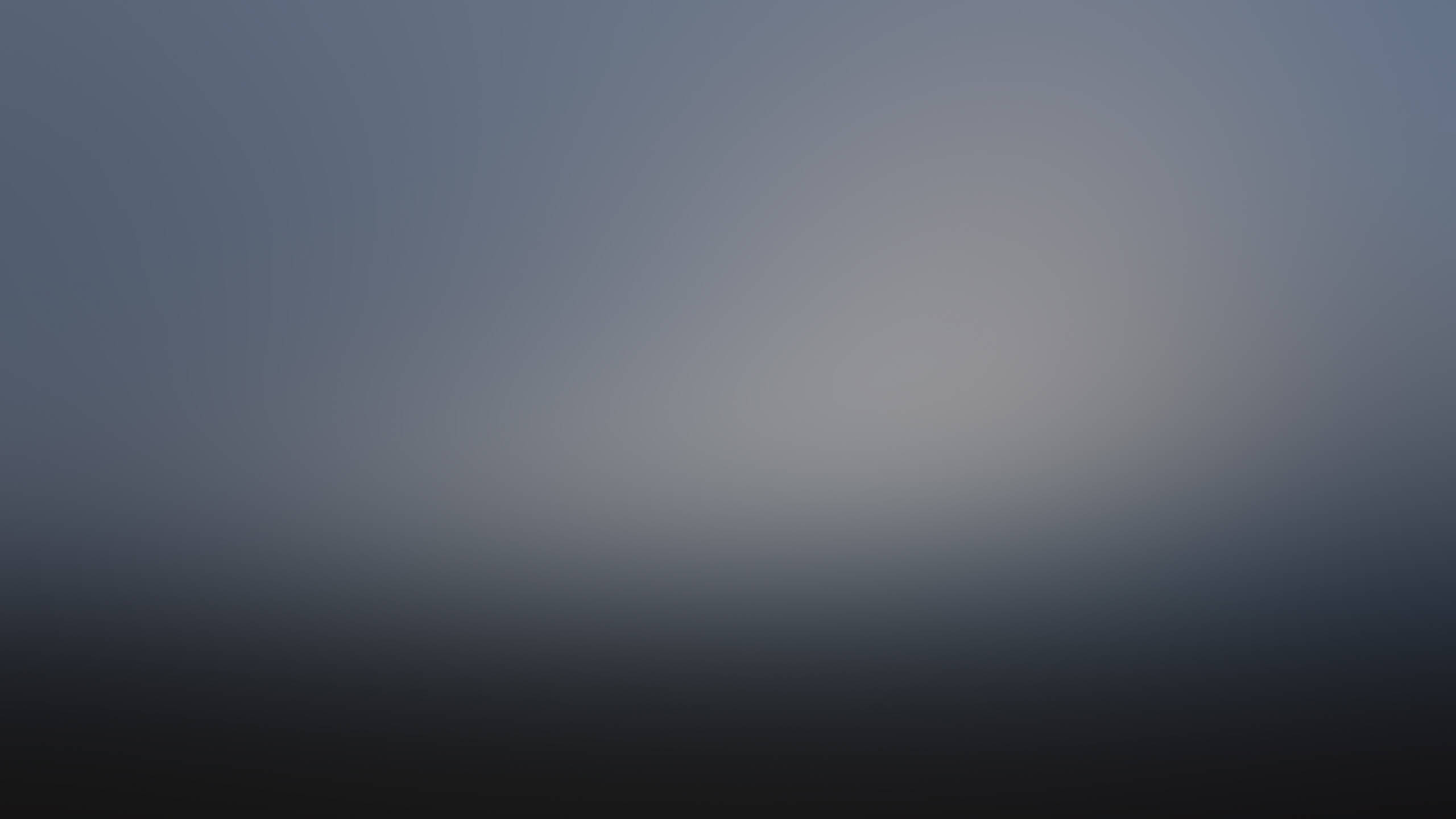 Blurry gradient image with a light gray center fading into darker blue and black tones toward the edges, creating a soft, abstract, and misty atmosphere.
