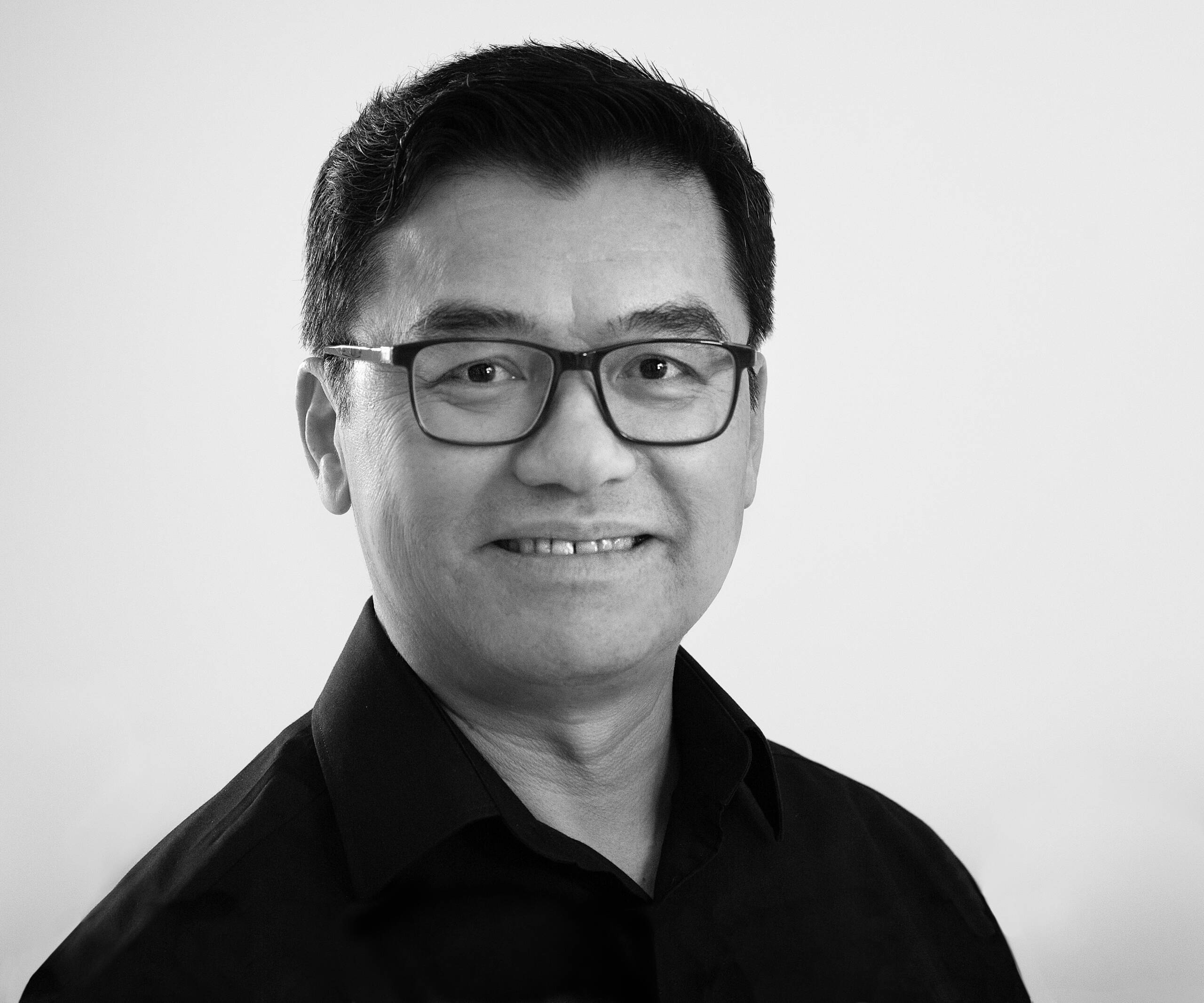 A man with short dark hair and glasses smiles at the camera. He is wearing a black collared shirt and is set against a plain, light background.
