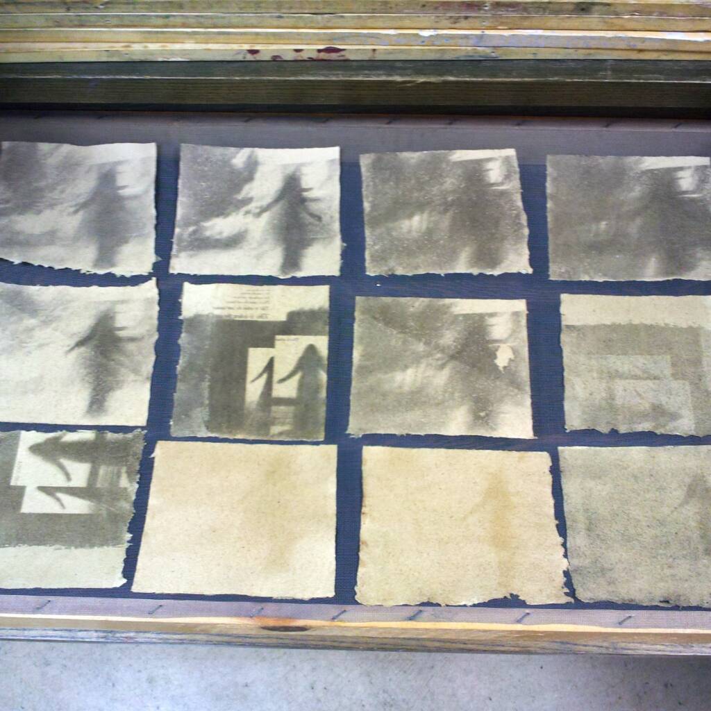 Twelve textured, square sheets of paper with abstract, shadowy grayscale and sepia patterns are laid out in a grid on a flat surface, possibly drying as part of an art or printmaking process.