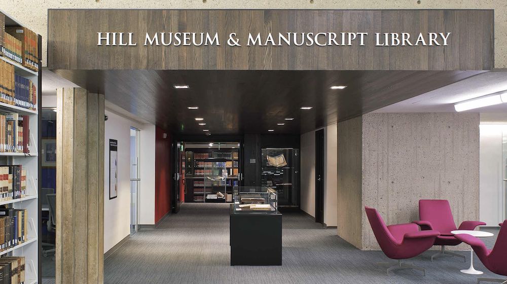Entrance to the Hill Museum & Manuscript Library, featuring glass display cases with books and manuscripts, bookshelves on the left, and modern pink chairs with a small table on the right.