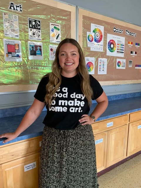 A woman stands smiling in a classroom, wearing a black T-shirt that reads "It's a good day to make some art." Behind her are bulletin boards displaying colorful art and color theory posters.