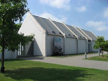 A modern, white building with a series of steep, gabled roofs stands among green grass and trees under a partly cloudy sky. A paved walkway leads to the entrance.