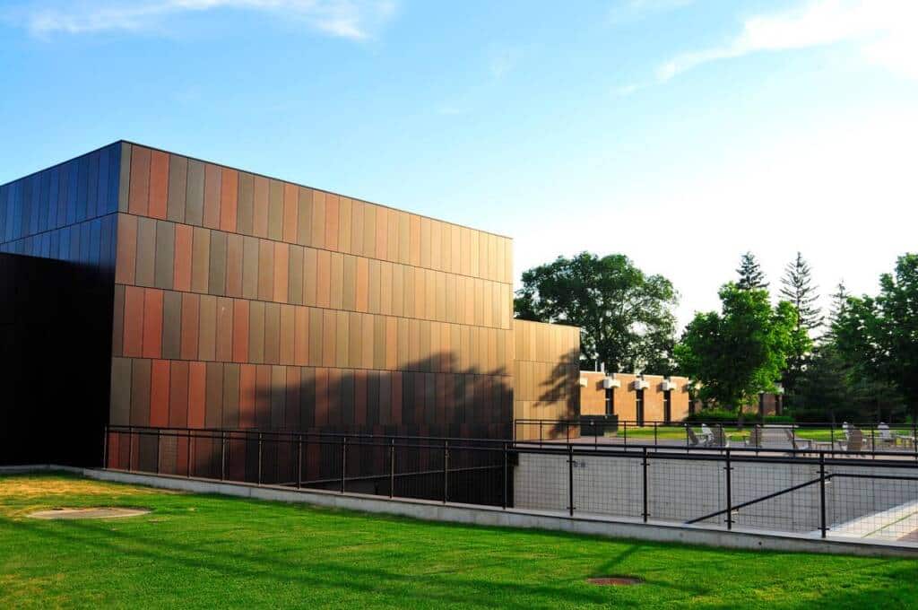 A modern, rectangular building with dark, reddish-brown panels stands beside a grassy lawn and a black metal fence. Trees and a clear blue sky are visible in the background.