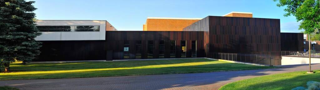 A modern building with dark brown and white exterior walls sits beside a green lawn and trees under a clear blue sky. A sidewalk curves in front of the structure.