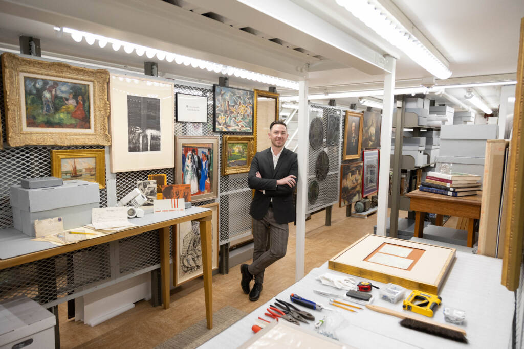 A person in a dark blazer stands in a well-lit room filled with framed artwork on the walls, tables with tools, storage boxes, and books, suggesting an art storage or conservation space.