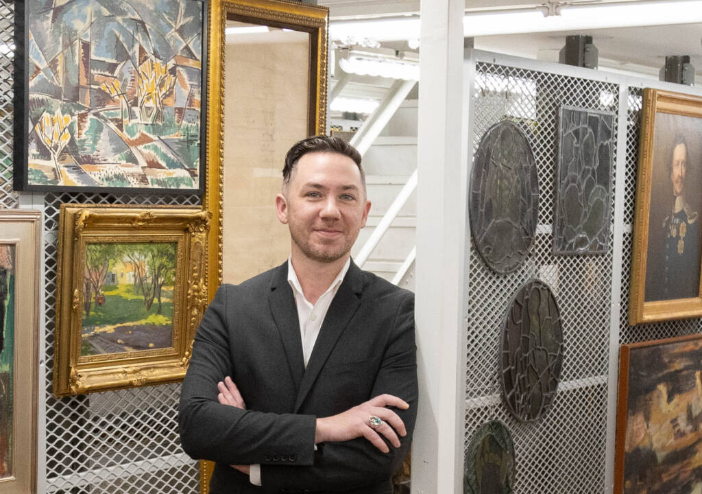 A person in a dark suit stands with arms crossed, smiling, in front of a wall displaying framed paintings and plaques in an art gallery or storage area.
