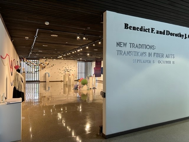 Gallery entrance with “New Traditions: Transitions in Fiber Arts” exhibition, featuring hanging and floor fiber art pieces in a well-lit space. Exhibition dates are September 5 to October 15.