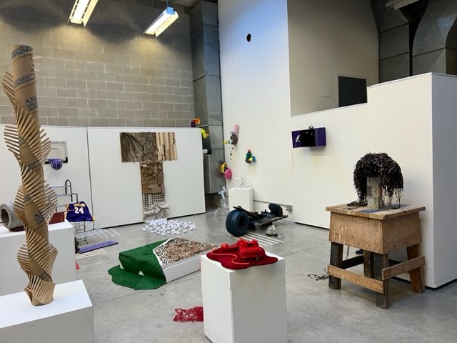 A contemporary art exhibit features eclectic sculptures made from everyday objects, including wood, cardboard, fabric, and metal, displayed throughout a white-walled gallery with concrete floors and exposed ceiling.