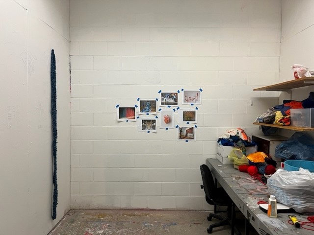 A small art studio with a long textured artwork on the left wall, several photos taped to the back wall, and shelves holding colorful fabric and yarn above a cluttered work desk with a black chair.