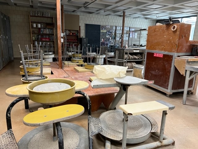 A ceramics studio with several pottery wheels and stools, shelves of supplies in the background, and a large kiln on the right. Stools are stacked on tables, and the room appears tidy and unoccupied.