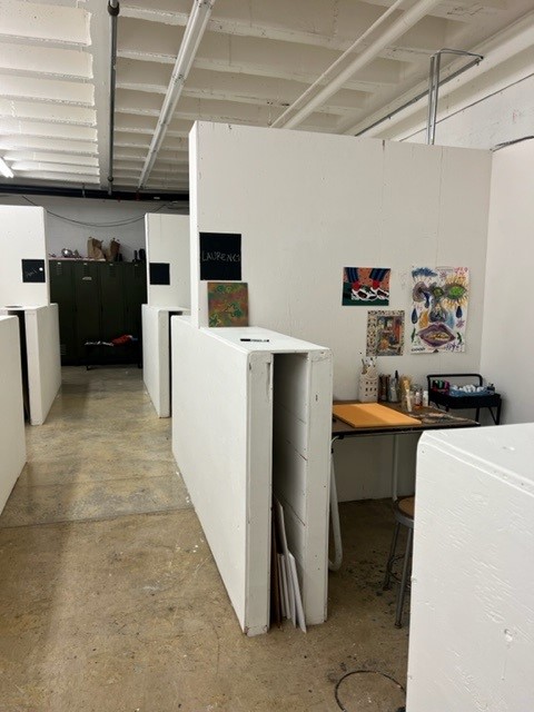 A studio space with white partition walls creating individual work areas. One area has a desk with art supplies, a stool, and colorful artwork and posters on the wall. The floor is concrete and the lighting is bright.