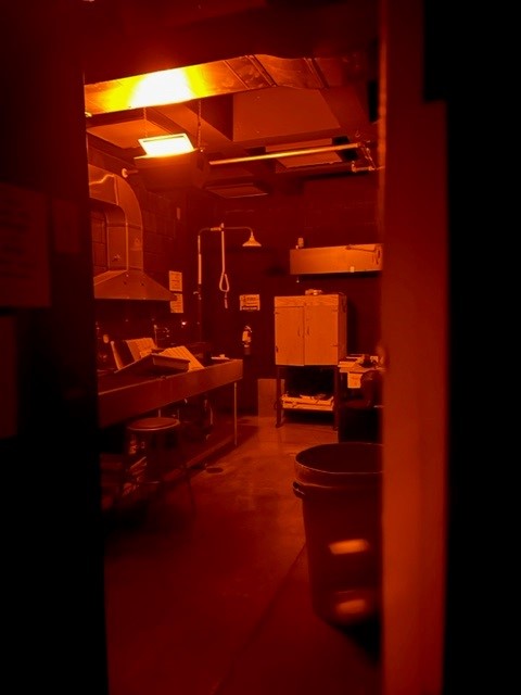 A dimly lit room with an intense red-orange glow, containing lab equipment, cabinets, a trash can, and various work surfaces scattered with items, suggesting a science lab viewed through a doorway.