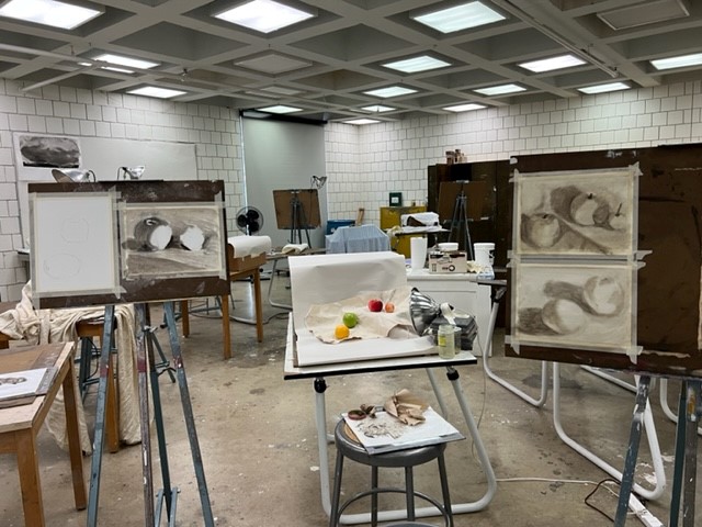 An art classroom with easels displaying charcoal still life drawings of fruit; tables hold various art supplies and real fruit arranged for reference, under bright ceiling lights.