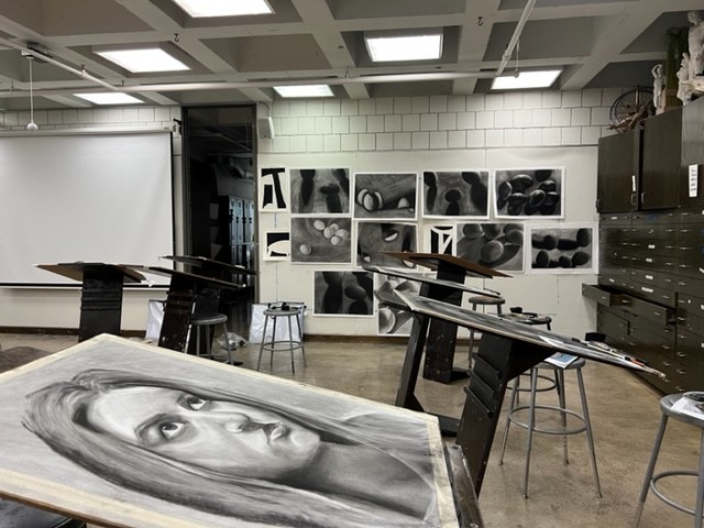 A classroom with drawing easels and stools is shown. Charcoal portraits and abstract artwork are displayed on the walls. A realistic charcoal drawing of a woman's face is on an easel in the foreground.