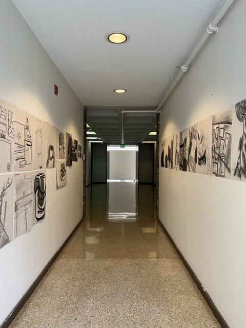 A hallway with white walls displaying black and white sketches on both sides, leading to a set of double doors and a staircase at the far end under bright ceiling lights.