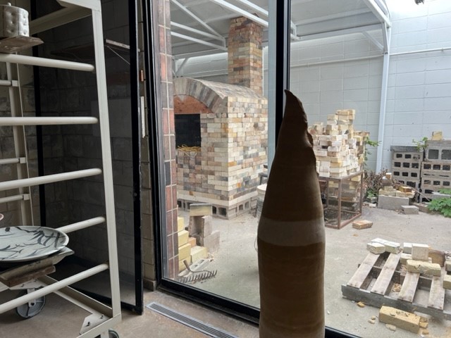 A view through a large window shows a brick kiln and stacks of bricks outside. Inside, there is a tall ceramic sculpture, a kiln shelf with pottery, and various materials scattered around the studio.