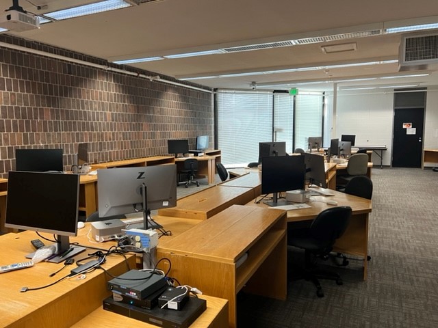 An empty office with several wooden desks arranged in rows, each with a computer, chair, and office equipment. The room has brick walls, carpeted floors, and large windows with closed blinds.
