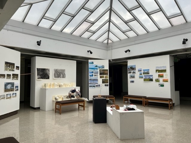 Art gallery with benches, sculptures on low pedestals, and walls displaying various landscape photographs and paintings. Natural light streams in from a large glass ceiling overhead.