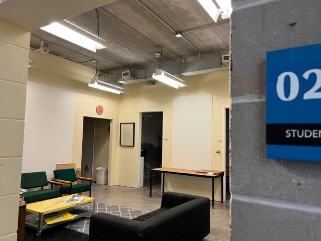 A student lounge with green and black seating, a coffee table with papers, and overhead fluorescent lights. The wall sign reads "021 Student" and there are open doors to adjacent rooms. The floor is concrete.