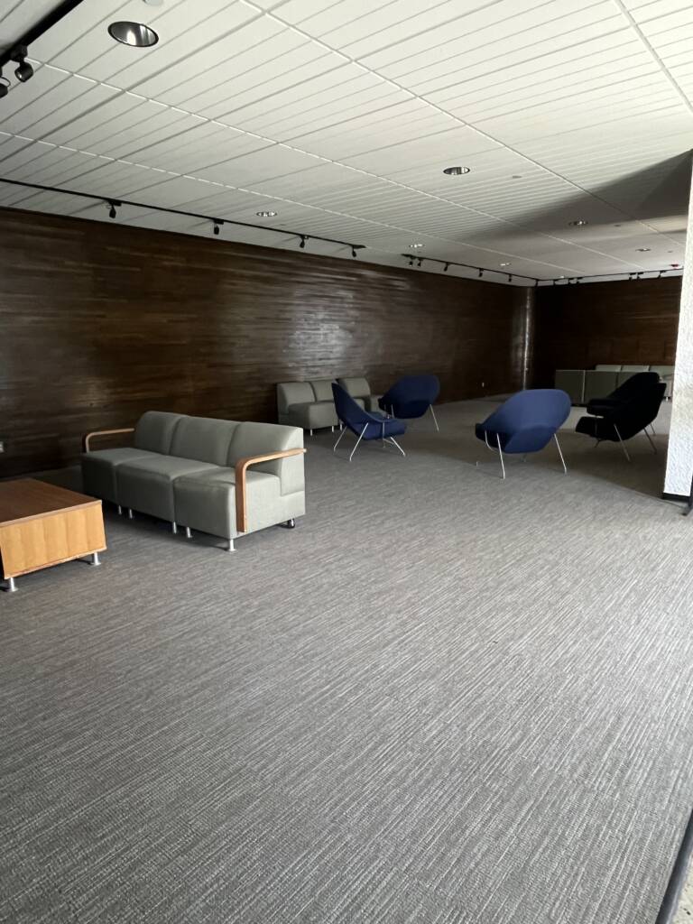 A modern lounge with gray sofas, blue armchairs, wooden tables, and a wood-paneled wall. The room is carpeted and well-lit with ceiling track lights, giving a spacious and minimalist feel.