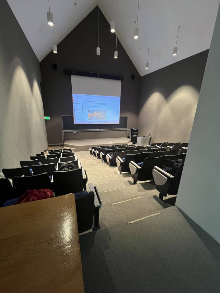A modern lecture hall with tiered seating, facing a large projection screen displaying a presentation. The room is empty, with soft lighting and white walls.