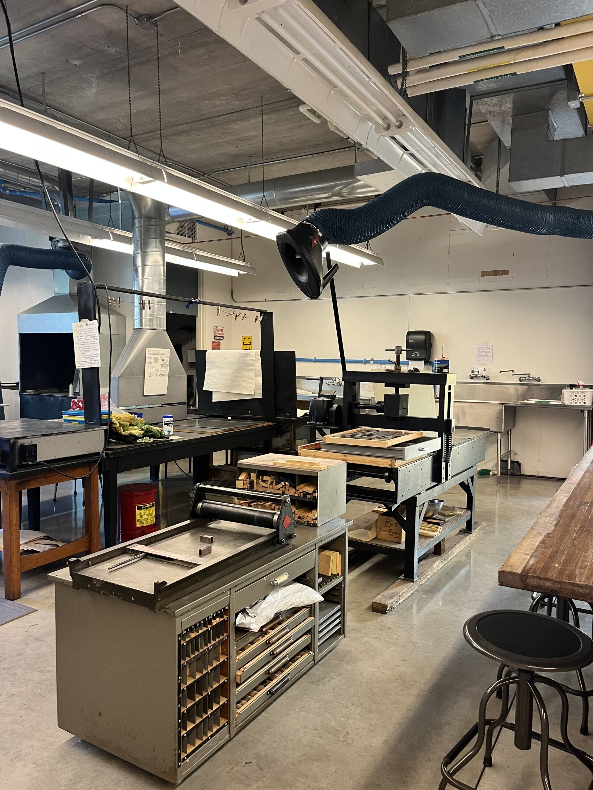 A well-lit workshop with various equipment, including a large printing press, worktables, stools, ventilation ducts, and tools. Papers and notes are posted on walls and equipment, and supplies are organized on shelves and in drawers.