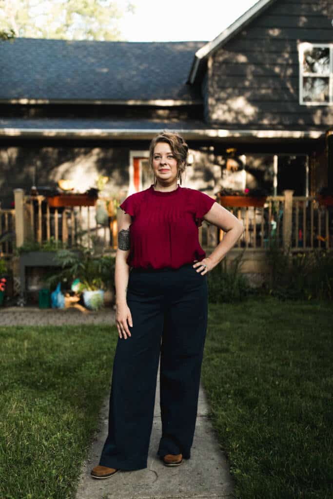 A woman with short hair stands confidently on a lawn in front of a dark-colored house, wearing a burgundy blouse and dark wide-leg pants, with one hand on her hip and a tattoo visible on her left arm.