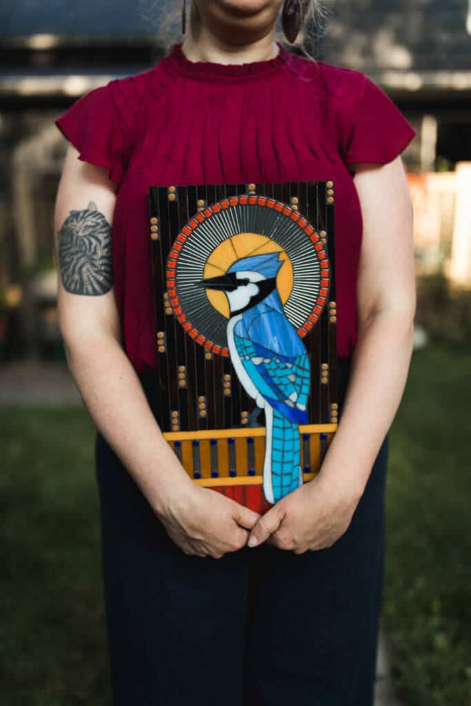 A person in a red top holds a colorful stained glass artwork depicting a blue jay with a circular, decorative background. The person has a tattoo on their upper arm and is standing outdoors.